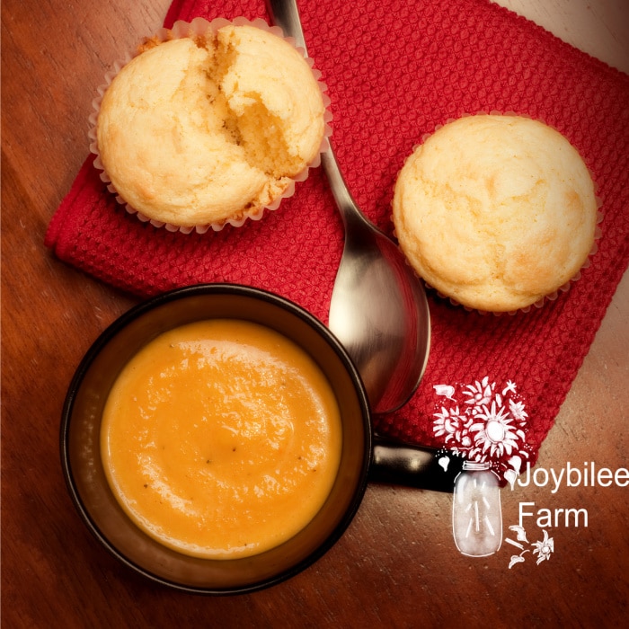 A mug full of creamy butternut squash bisque with corn muffins on a wooden table.